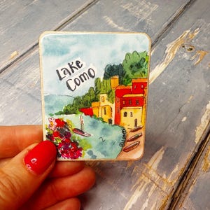 May include: A watercolour painting of a lakeside village with colourful buildings and boats. The text "Lake Como" is written in black cursive at the top of the image.
