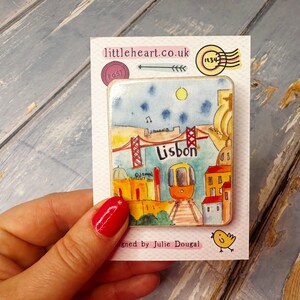 May include: A colorful ceramic magnet featuring a cartoon illustration of Lisbon, Portugal. The magnet depicts a red bridge, a yellow tram, and buildings in a watercolor style. The text "Lisbon" is written in red letters on the magnet.