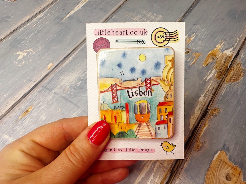 May include: A colorful ceramic magnet featuring a cartoon illustration of Lisbon, Portugal. The magnet depicts a red bridge, a yellow tram, and buildings in a watercolor style. The text "Lisbon" is written in red letters on the magnet.