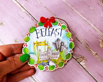 May include: A round, wooden ornament with a white background and a green holly wreath border. The ornament features a watercolor illustration of landmarks in Belfast, Northern Ireland, including the Albert Memorial Clock, the Grand Opera House, and the Titanic Belfast. The text "BELFAST" is written in black across the top of the ornament.