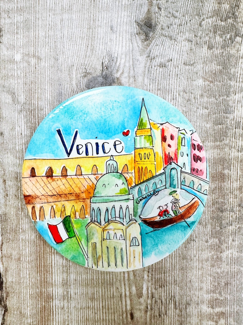 May include: A round ceramic plate with a watercolor painting of Venice, Italy. The plate features a blue sky, a yellow building with arches, a green domed building, a tall red and yellow tower, and a gondola with two people in it. The words "Venice" are written in black on the plate.