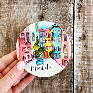 Pode incluir: Uma base para copos redonda com uma pintura em aquarela de uma cena de rua em Istambul. Os edifícios são pintados em cores vibrantes, com um carro azul na rua. A palavra "Istambul" está escrita abaixo.