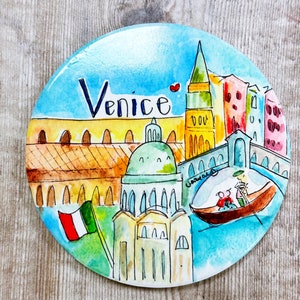 May include: A round, blue and white ceramic magnet with a watercolor painting of Venice, Italy. The painting features a gondola, a church with a dome, a tall building with a spire, and a bridge. The words "Venice" are written in black at the top of the magnet.
