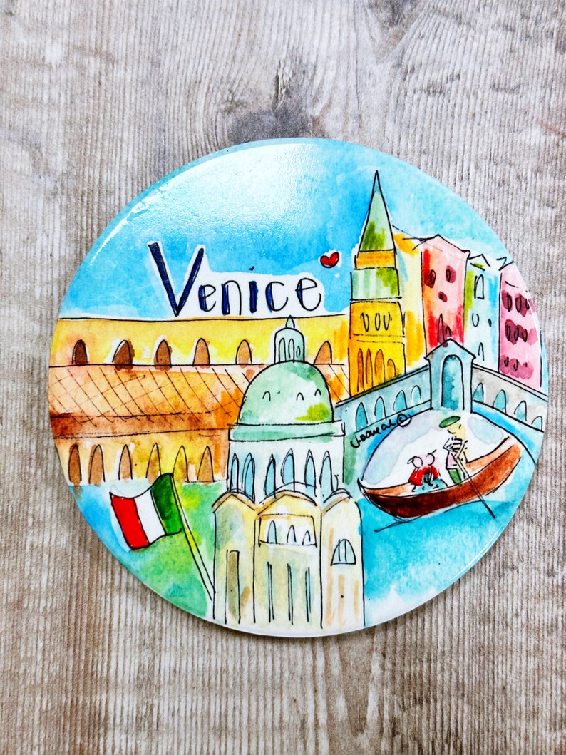 May include: A round, blue and white ceramic magnet with a watercolor painting of Venice, Italy. The painting features a gondola, a church with a dome, a tall building with a spire, and a bridge. The words "Venice" are written in black at the top of the magnet.