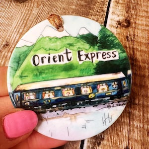 May include: A round, white ceramic ornament featuring a watercolour painting of a train and mountains. The train is blue and black with windows. The mountains are green and the text "Orient Express" is written in black on a white banner.