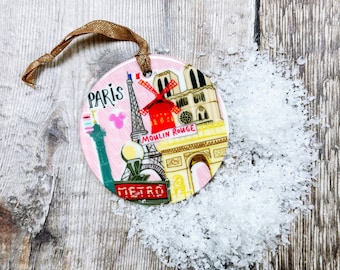 May include: A pink and white round ceramic ornament with a depiction of iconic Parisian landmarks, including the Eiffel Tower, the Arc de Triomphe, and the Moulin Rouge. The ornament has a gold ribbon for hanging and the text "Paris" and "Metro" are visible.