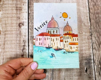 May include: Watercolor painting of Venice featuring buildings with domes, boats on turquoise water, and a yellow sun. The word "Venice" is written in black script. The artwork is on a rectangular card.
