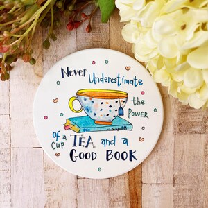 May include: White ceramic coaster with a colorful illustration of a teacup and a book. The text reads "Never Underestimate the Power of a Cup of Tea and a Good Book".