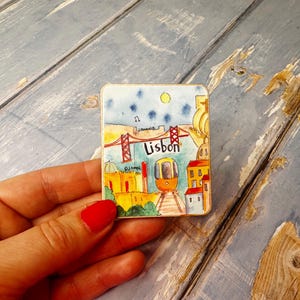 May include: A colorful ceramic magnet featuring a cartoon illustration of Lisbon, Portugal. The magnet depicts a red bridge, a yellow tram, and buildings with red roofs. The word "Lisbon" is written in black letters on the magnet.