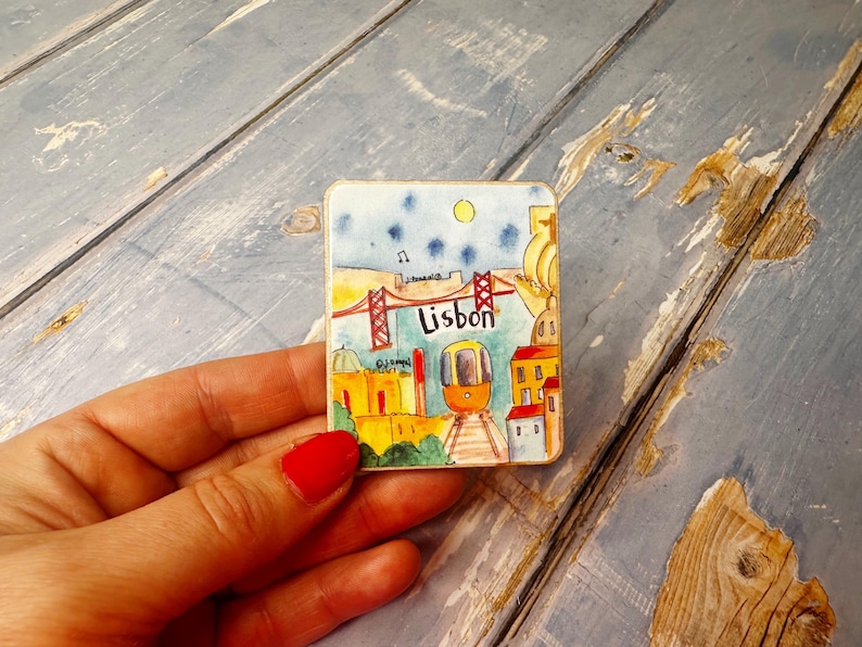 May include: A colorful ceramic magnet featuring a cartoon illustration of Lisbon, Portugal. The magnet depicts a red bridge, a yellow tram, and buildings with red roofs. The word "Lisbon" is written in black letters on the magnet.