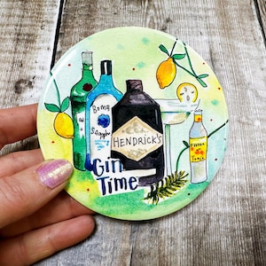 May include: A round, green and white coaster with a watercolor illustration of gin bottles, lemons, and a cocktail glass. The text "Gin Time" is written in black lettering.