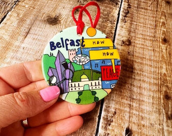 May include: A round ceramic ornament with a colorful illustration of Belfast, Northern Ireland. The ornament features landmarks, text, and a red ribbon for hanging.