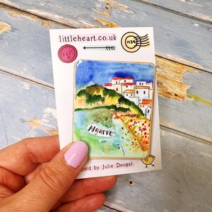 May include: A watercolor painting of Algarve, Portugal, on a postcard. The artwork depicts a coastal scene with white buildings, green hills, and blue water under a blue sky. The postcard has the text "littleheart.co.uk".