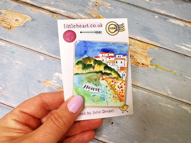 May include: A watercolor painting of Algarve, Portugal, on a postcard. The artwork depicts a coastal scene with white buildings, green hills, and blue water under a blue sky. The postcard has the text "littleheart.co.uk".