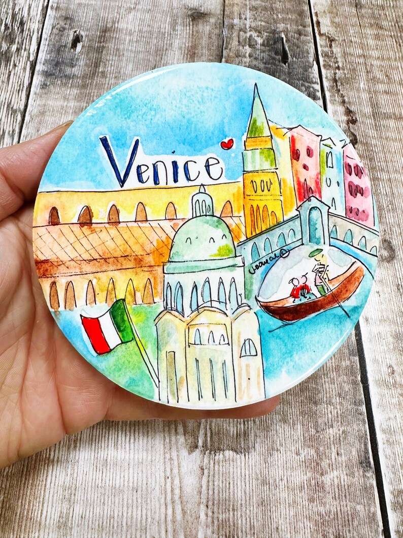 May include: A round ceramic plate with a watercolor painting of Venice, Italy. The plate features a blue sky, a red heart, a yellow building with a green dome, a tall yellow tower, and a gondola with two people in it. The words "Venice" are written in black on the plate.