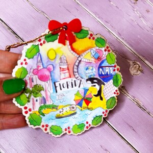 May include: A round, wooden ornament with a colorful illustration of Florida landmarks, including a lighthouse, a palm tree, a Mickey Mouse head, and the words "Florida" and "NASA". The ornament is decorated with red berries and green holly leaves.