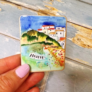 May include: A watercolor painting of the Algarve coast, featuring white buildings with red roofs, a green hillside, and a beach. The word "Algarve" is written in cursive. The artwork is on a small, rectangular piece.
