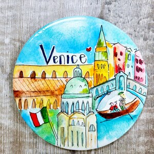 May include: A round, colorful magnet with a watercolor painting of Venice, Italy. The magnet features a gondola with two people, a tall building with a red roof, and the word "Venice" written in black.