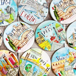 May include: Set of ten colorful round coasters featuring hand-painted illustrations of famous landmarks from cities around the world, including Paris, London, Rome, New York, Dublin, Belfast, Barcelona, and Venice. Each coaster has the city name written in a whimsical font.