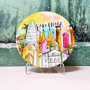 May include: A round ceramic plate with a watercolor painting of the Barcelona skyline. The plate features iconic buildings like the Sagrada Familia and the Agbar Tower. The plate is on a clear acrylic stand.