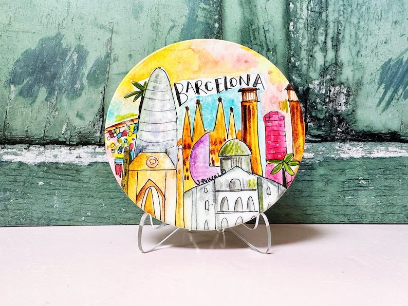 May include: A round ceramic plate with a watercolor painting of the Barcelona skyline. The plate features iconic buildings like the Sagrada Familia and the Agbar Tower. The plate is on a clear acrylic stand.