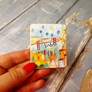 May include: A colorful magnet featuring a cartoon illustration of Lisbon, Portugal. The magnet depicts a red bridge, a yellow tram, and buildings in a watercolor style. The word "Lisbon" is written in black letters.