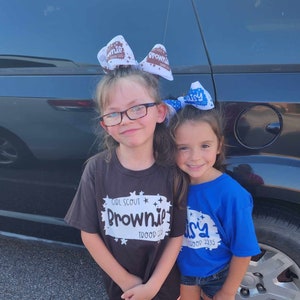 May include: Two young girls wearing Girl Scout uniforms. The girl on the left is wearing a black shirt with the words "Girl Scout Brownie Troop 2233" printed on it. The girl on the right is wearing a blue shirt with the words "Daisy Troop 2233" printed on it. Both girls are wearing bows in their hair.
