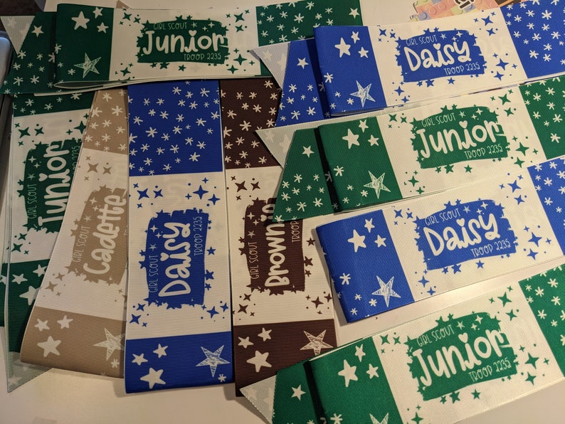 May include: A collection of Girl Scout sashes with different troop numbers and levels, including Junior, Daisy, Cadette, and Brownie. The sashes are decorated with white stars on a variety of colored backgrounds, including green, blue, brown, and beige.