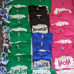 May include: A collection of green, pink, blue, black, and beige t-shirts with white text and star designs. The text on the shirts includes "Girl Scout", "Junior", "Brownie", "Daisy", "Leader", "Cadette", and "Mom". The shirts also include the troop number "2235".