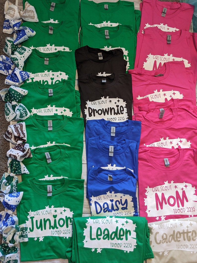 May include: A collection of green, pink, blue, black, and beige t-shirts with white text and star designs. The text on the shirts includes "Girl Scout", "Junior", "Brownie", "Daisy", "Leader", "Cadette", and "Mom". The shirts also include the troop number "2235".