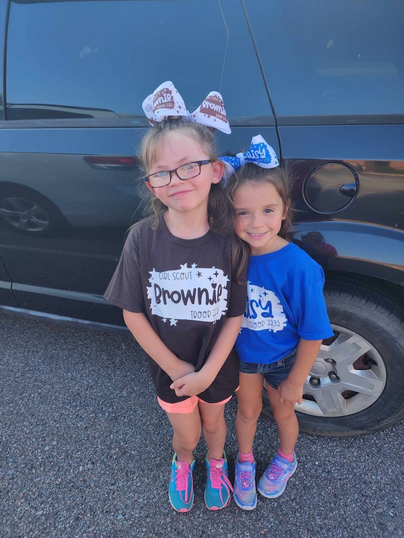 May include: Two young girls wearing Girl Scout uniforms. The girl on the left is wearing a black shirt with the words "Girl Scout Brownie Troop 2233" printed on it. The girl on the right is wearing a blue shirt with the words "Daisy Troop 2233" printed on it. Both girls are wearing bows in their hair.