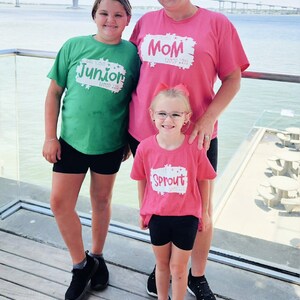 May include: Three people wearing pink and green t-shirts with the text "Mom Troop 2355", "Junior Troop 2355", and "Sprout Troop 2355" printed on them. They are standing on a wooden deck with a body of water in the background.
