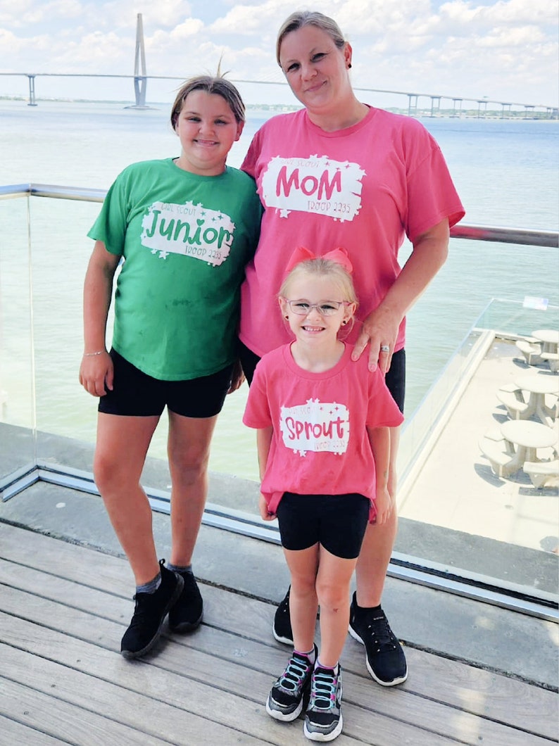 May include: Three people wearing pink and green t-shirts with the text "Mom Troop 2355", "Junior Troop 2355", and "Sprout Troop 2355" printed on them. They are standing on a wooden deck with a body of water in the background.