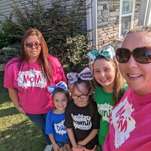 May include: A group of five people wearing pink, green, blue, and black t-shirts with the text "Girl Scout" and the troop number "2235". The shirts have different designs, including a star, a brownie, and a junior. The people are smiling and looking at the camera.