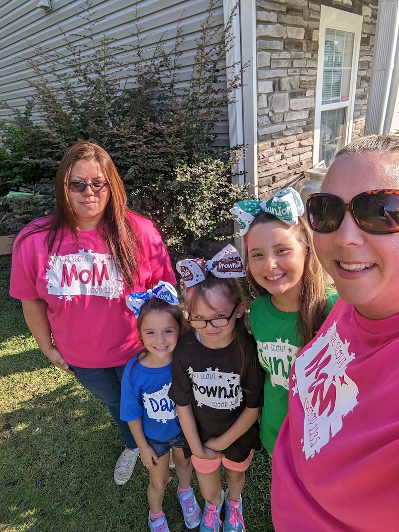 May include: A group of five people wearing pink, green, blue, and black t-shirts with the text "Girl Scout" and the troop number "2235". The shirts have different designs, including a star, a brownie, and a junior. The people are smiling and looking at the camera.