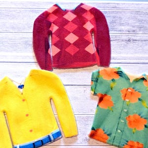 Può includere: Tre capi di abbigliamento in miniatura in tessuto: un maglione bordeaux a rombi, un cardigan giallo con dettaglio cintura blu e una camicia hawaiana turchese con motivi floreali arancioni. Gli articoli sono disposti su una superficie chiara con venature del legno.