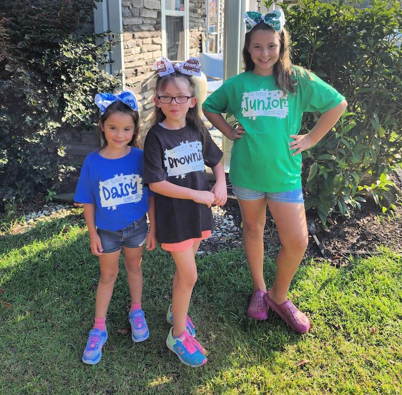 May include: Three girls wearing Girl Scout t-shirts with their troop level printed on them. The girl on the left is wearing a blue shirt with "Daisy" printed on it. The girl in the middle is wearing a black shirt with "Brownie" printed on it. The girl on the right is wearing a green shirt with "Junior" printed on it.