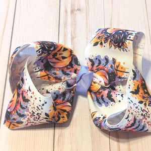 Könnte beinhalten: Eine große Haarschleife mit Halloween-Motiv. Die Schleife hat eine cremefarbene Basis mit orangefarbenen, schwarzen und lila Mustern, darunter Fledermäuse, Kürbisse und florale Motive. Ein hellblaues Band sichert die Schleife.