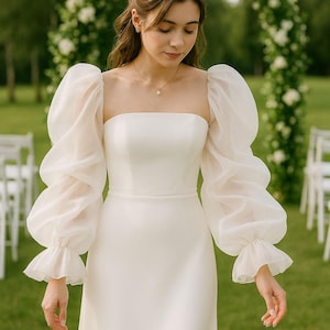 May include: A white wedding dress with a square neckline and sheer, puffy sleeves. The dress has a fitted bodice and a flowing skirt. The sleeves are long and end in ruffled cuffs. The background includes a floral arch and green grass.