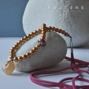May include: A gold-coloured beaded necklace with a round, light orange pendant. The necklace features a dark red bead and a matching dark red suede cord. The necklace is displayed on a white stone with the text "FALCONE JEWELLERY ART GALLERY" in the background.