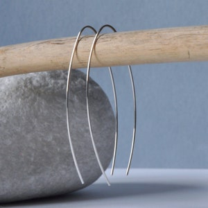 Può includere: Tre orecchini a cerchio d'argento, ognuno con un design a filo sottile e curvo, sono esposti su uno sfondo blu chiaro. Gli orecchini sono appoggiati su un pezzo di legno galleggiante e una pietra grigia.