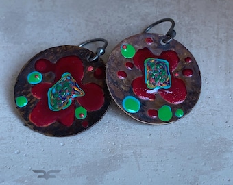 May include: A pair of round, dark-toned metal earrings featuring a textured surface with glossy red enamel accents, bright green circular dots, and a central multicolored abstract swirl. Each earring hangs from a dark metal hook.