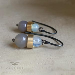 May include: A pair of drop earrings featuring a large grey spherical bead, a central cylindrical gold-toned metal spacer, and a smaller translucent pale blue bead. The earrings are attached to dark grey metal hooks with the text FALCONE below.