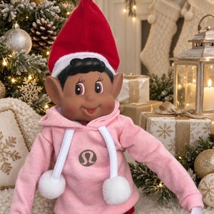 May include: A dark-skinned elf doll wearing a red pointed hat with a white trim and a light pink hooded sweatshirt featuring a circular brown logo on the chest, white drawstrings with pom-poms, and red and white striped leggings on its legs.