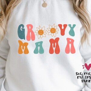 May include: A white sweatshirt with the text "Groovy Mama" in colorful retro-style letters. Two yellow sunflowers with smiling faces are above the text. The text "SVG, PNG, JPG, DXF, EPS, PDF BUNDLE" is at the bottom of the image.