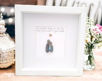May include: A white framed picture with a quote that reads "the smallest things can take up most of the room in your heart". The quote is written in black ink. There is a small gold heart above the quote. Below the quote is a family of three made of gray and blue stones. The family is made up of two adults and a child. The picture is sitting on a wooden surface.