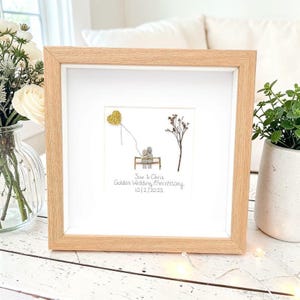 以下が含まれることがあります： 明るい木製フレームに入った額装されたアートワーク。アートワークは、ベンチに座っているカップル、金のグリッターハートの風船、木を特徴としています。「Sue & Chris Golden Wedding Anniversary 10/2/2023」と書かれています。