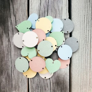 May include: A collection of wooden shapes in various pastel colours, including circles, hearts, and teardrop shapes. The shapes have small holes punched in them, suggesting they are for crafting or decorating.