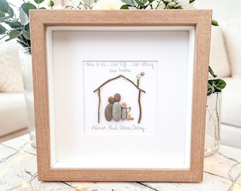 May include: A framed artwork featuring a family and home design made of pebbles. The artwork is in a light brown wooden frame with a white mat. The text "This is us... our life... our story... our home" is written above the family illustration.