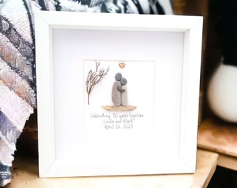 May include: A white framed picture with a stone art piece of a couple embracing. The art piece is on a small piece of driftwood with a small branch above them. The text below reads "Celebrating 50 years together Linda and Mark April 23, 2023".
